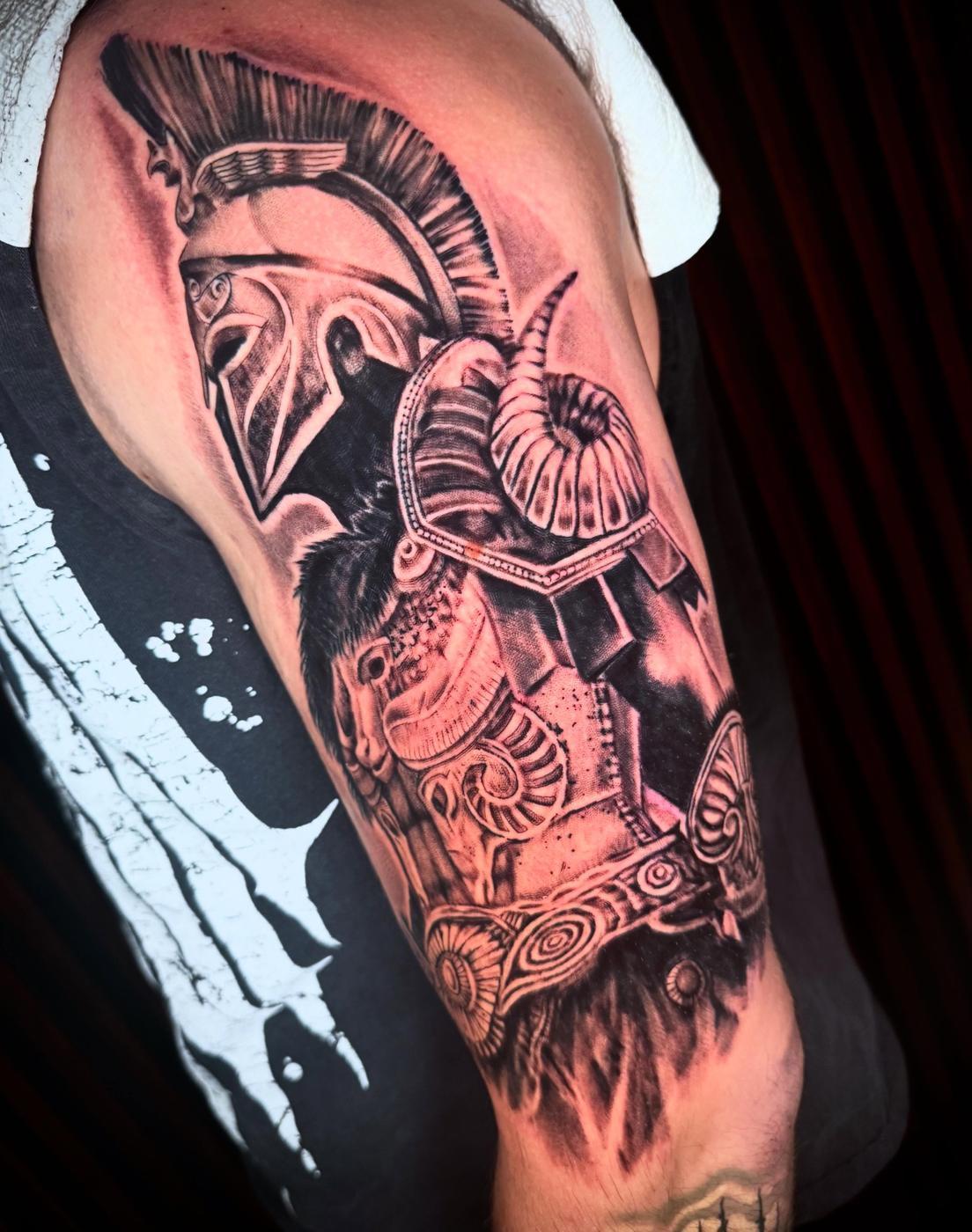 A black and gray realism Spartan warrior in a plumed helmet, on an upper arm.