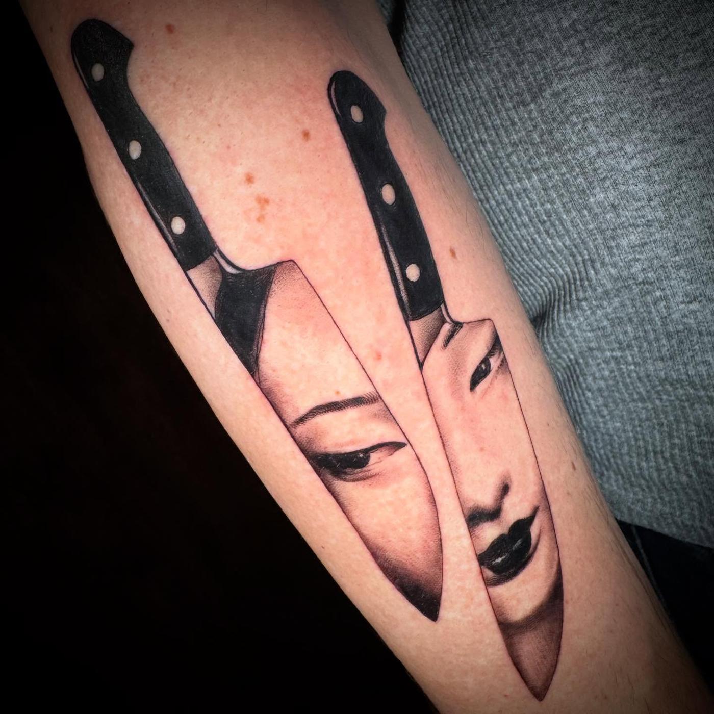 Two black and gray kitchen knives with a woman's face reflected across both blades, on a forearm.