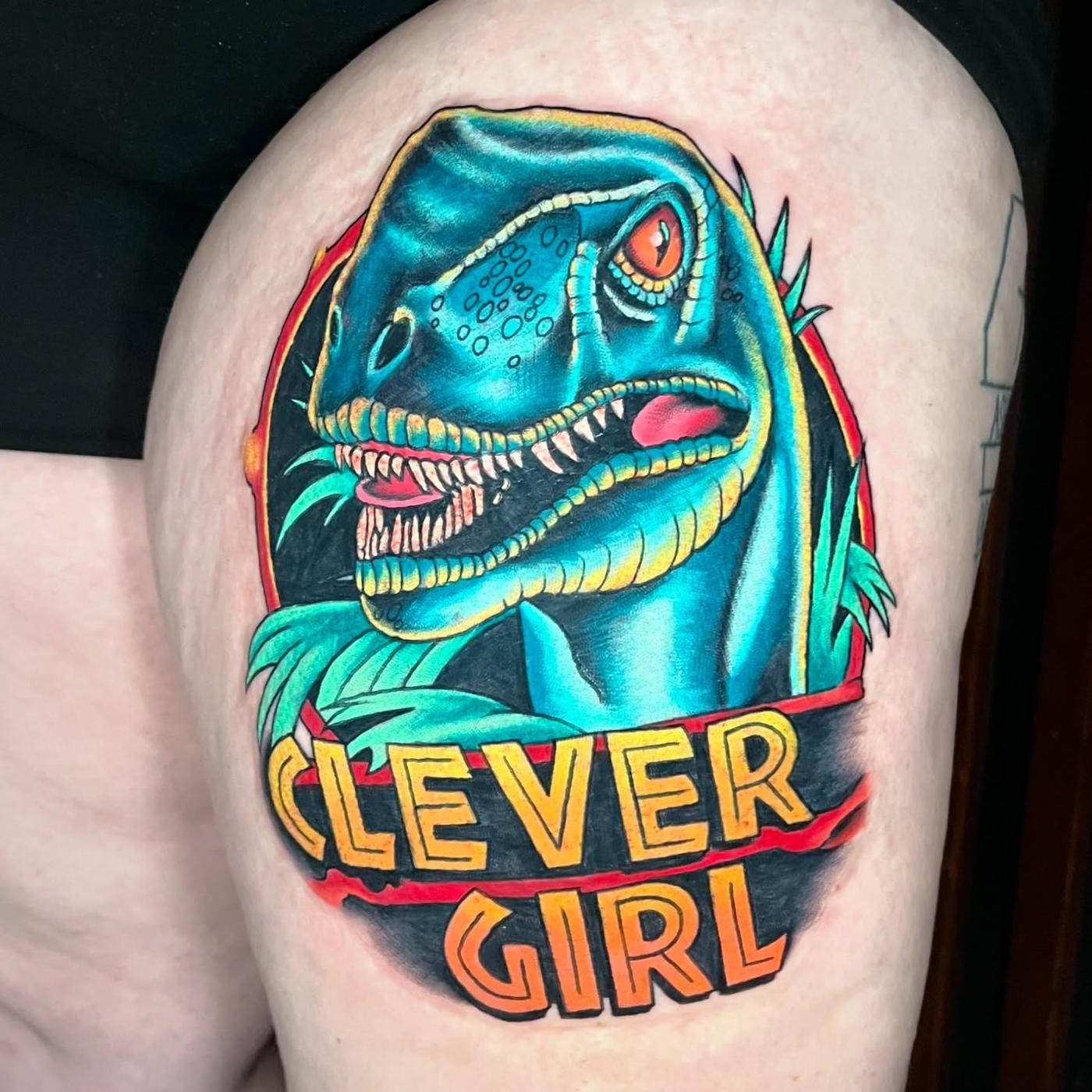A neo-traditional raptor head above the words Clever Girl, on a thigh.
