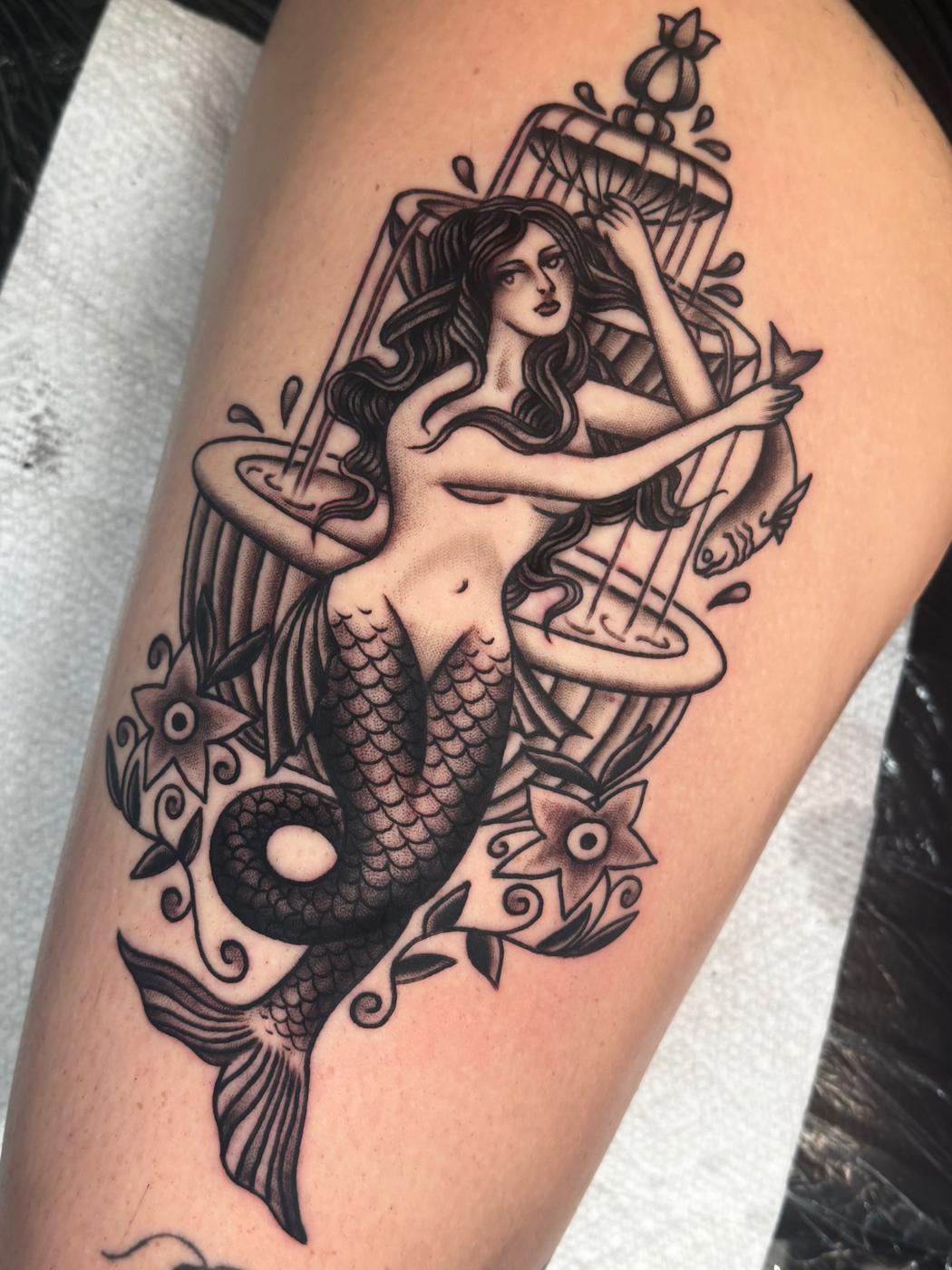 A black and gray traditional mermaid in front of a birdcage, on a thigh.