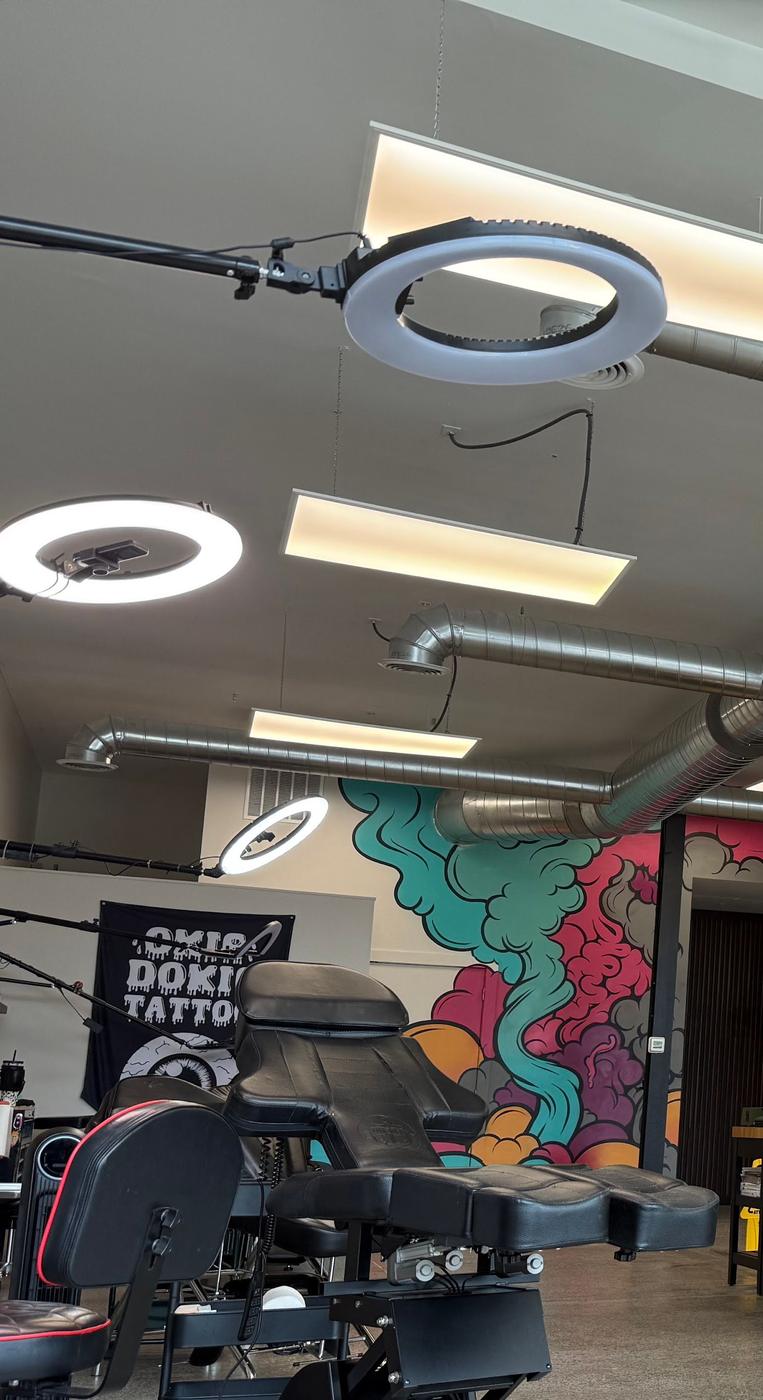 Inside the shop: a tattoo chair and ring lights under exposed ductwork, with a turquoise and pink mural along the back wall and an Okie Dokie Tattoo banner.