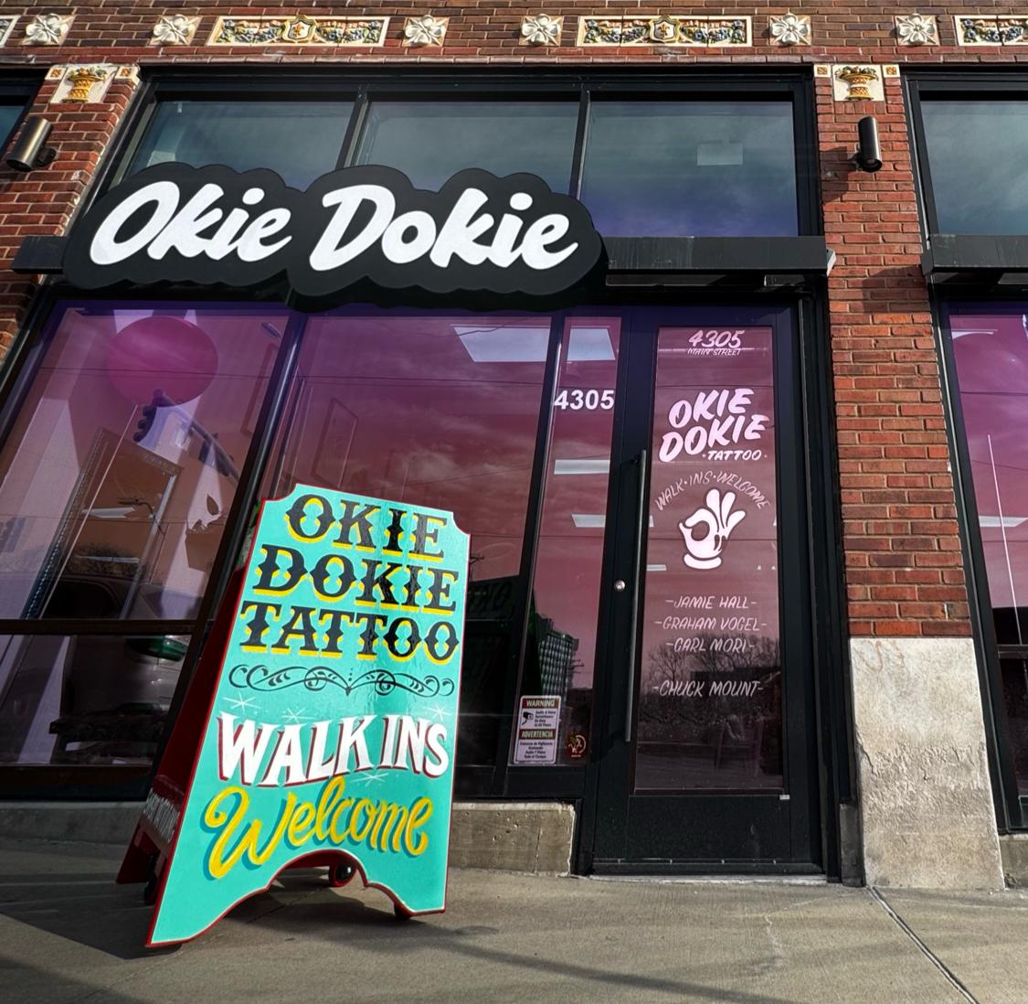 The Okie Dokie storefront on Main Street: a black cloud-shaped sign in white script above the window, and a hand-painted board on the sidewalk reading Okie Dokie Tattoo, Walk Ins Welcome.